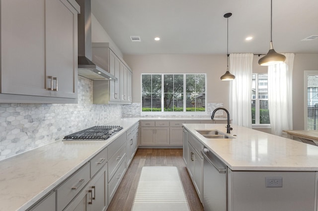 kitchen with pendant lighting, sink, gray cabinets, a kitchen island with sink, and wall chimney exhaust hood