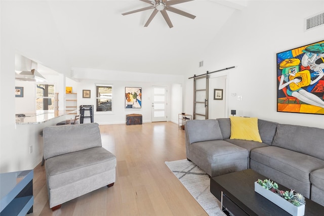 living room with ceiling fan, a barn door, high vaulted ceiling, and light wood-type flooring