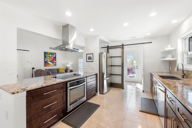 kitchen with sink, light stone counters, island range hood, stainless steel appliances, and a barn door