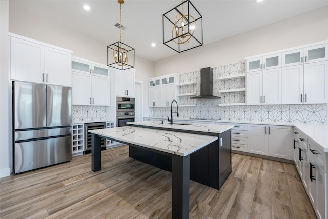 kitchen with pendant lighting, appliances with stainless steel finishes, a kitchen island with sink, white cabinets, and wall chimney exhaust hood