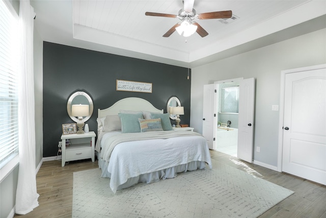 bedroom with ensuite bathroom, light wood-type flooring, a tray ceiling, ceiling fan, and multiple windows