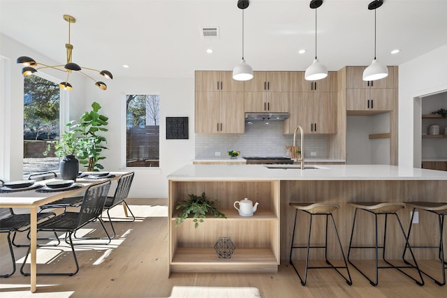 kitchen featuring sink, tasteful backsplash, hanging light fixtures, light wood-type flooring, and a kitchen island with sink