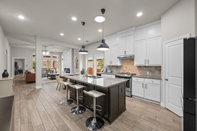 kitchen with stainless steel gas stove, light stone counters, white cabinetry, refrigerator, and a center island with sink