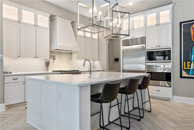 kitchen featuring built in appliances, an island with sink, white cabinets, and premium range hood