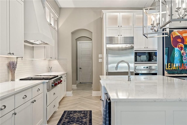 kitchen with hanging light fixtures, custom range hood, built in appliances, and a kitchen island with sink