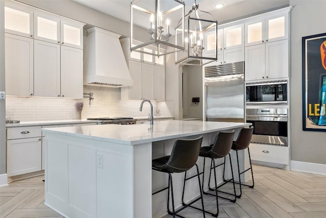 107 Bisset Ct, Austin, TX 78738 - kitchen with built in appliances, custom exhaust hood, tasteful backsplash, white cabinetry, and an island with sink
