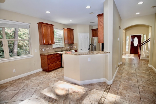 kitchen with light tile patterned floors, stainless steel fridge, kitchen peninsula, light stone countertops, and backsplash