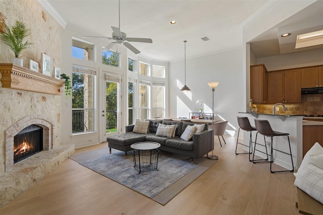 living room with a stone fireplace, a towering ceiling, ceiling fan, crown molding, and light hardwood / wood-style flooring