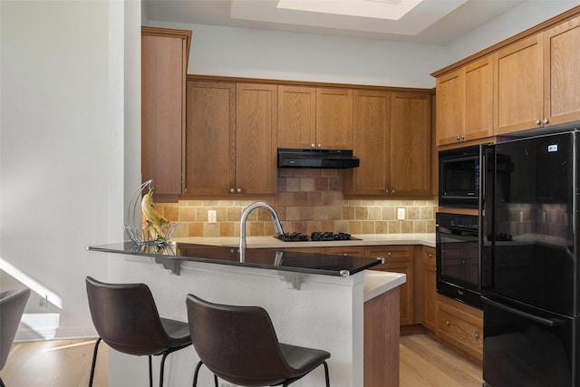 kitchen featuring a breakfast bar, black appliances, decorative backsplash, kitchen peninsula, and light wood-type flooring