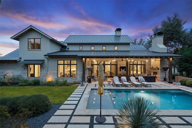 back of property with a patio, stone siding, a chimney, a standing seam roof, and stucco siding