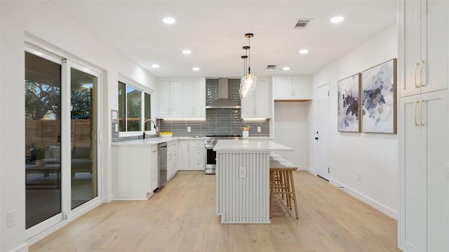 kitchen featuring appliances with stainless steel finishes, white cabinets, hanging light fixtures, a center island, and wall chimney range hood