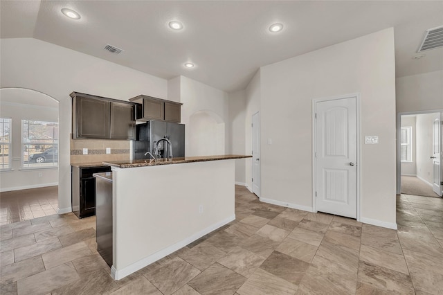 kitchen featuring vaulted ceiling, an island with sink, backsplash, dark brown cabinetry, and black refrigerator with ice dispenser