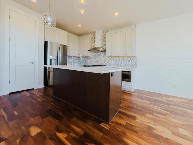 kitchen featuring stainless steel refrigerator with ice dispenser, decorative light fixtures, a center island with sink, dark hardwood / wood-style floors, and wall chimney range hood