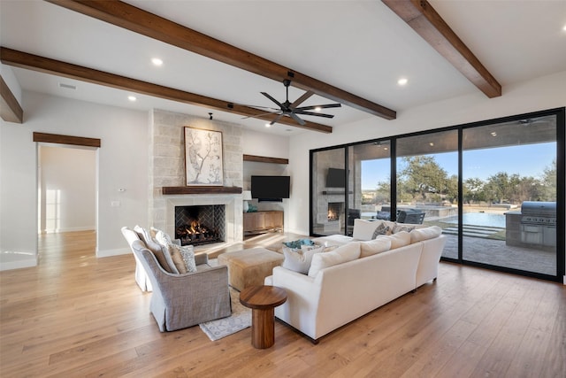 living room featuring ceiling fan, a stone fireplace, light hardwood / wood-style floors, and beam ceiling