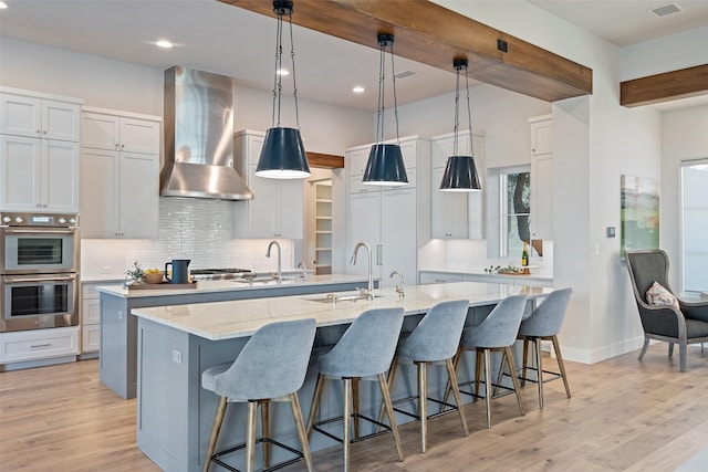 kitchen with white cabinetry, an island with sink, pendant lighting, and wall chimney exhaust hood