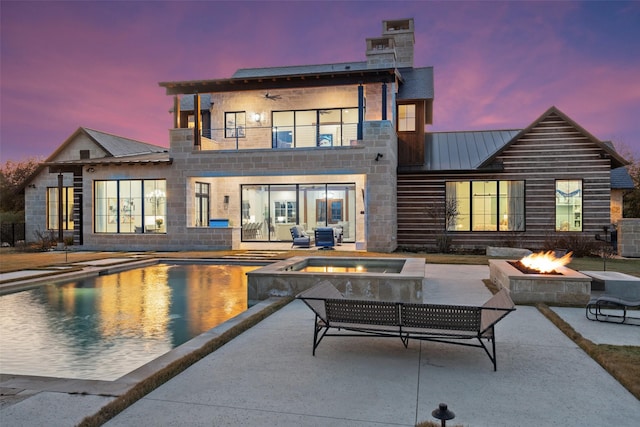 back house at dusk featuring a pool with hot tub, a fire pit, a patio, and a balcony