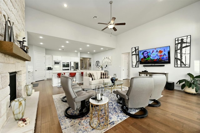 living room featuring recessed lighting, a towering ceiling, dark wood-type flooring, a ceiling fan, and a stone fireplace