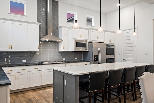 kitchen with wall chimney range hood, appliances with stainless steel finishes, hanging light fixtures, white cabinets, and a kitchen island