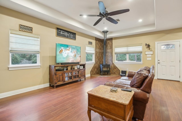 living room with ceiling fan, a raised ceiling, hardwood / wood-style floors, and a wood stove