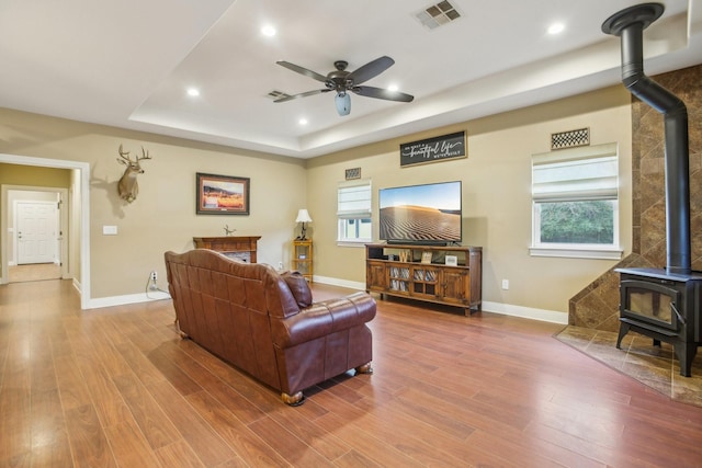 living room with wood-type flooring, a raised ceiling, ceiling fan, and a wood stove