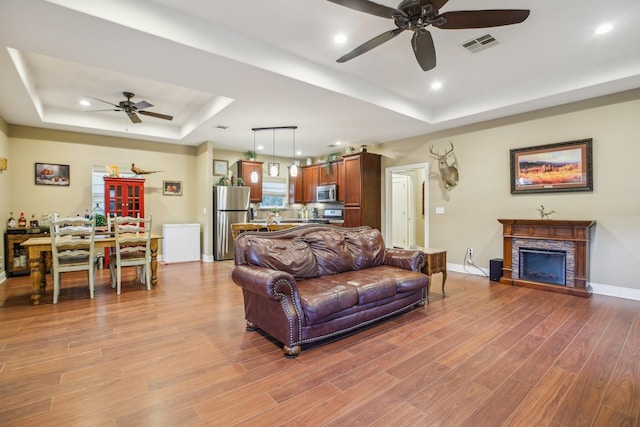 living room featuring a stone fireplace, light hardwood / wood-style flooring, ceiling fan, and a tray ceiling