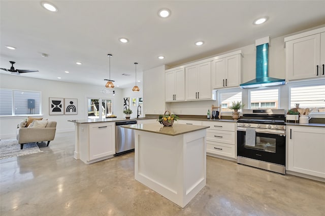 kitchen featuring dark countertops, wall chimney exhaust hood, appliances with stainless steel finishes, open floor plan, and decorative light fixtures