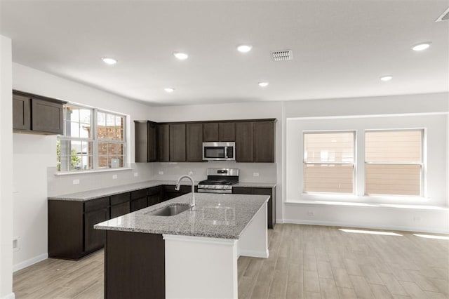 kitchen featuring an island with sink, light stone counters, a sink, appliances with stainless steel finishes, and light wood-style flooring