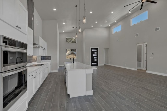 kitchen featuring double oven, decorative light fixtures, white cabinets, a high ceiling, and a kitchen island with sink