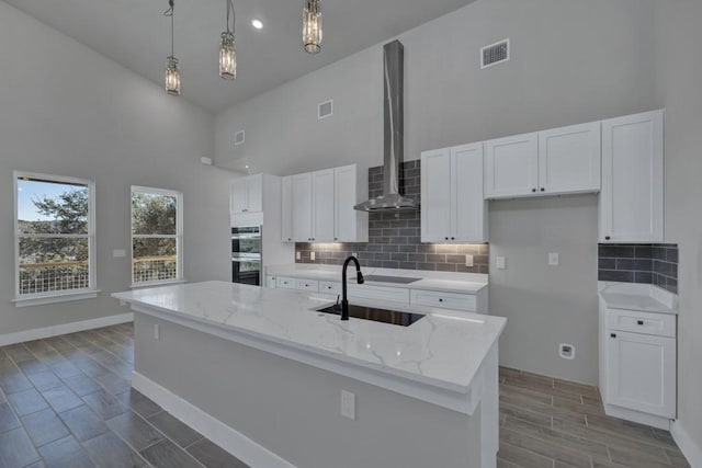 kitchen with pendant lighting, wall chimney range hood, a kitchen island with sink, and white cabinets