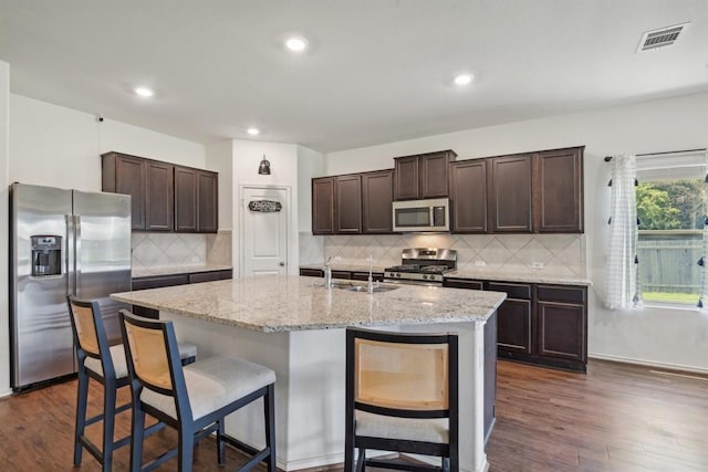 kitchen with appliances with stainless steel finishes, tasteful backsplash, a kitchen breakfast bar, a kitchen island with sink, and dark brown cabinets