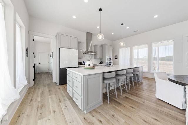 kitchen featuring wall chimney exhaust hood, gray cabinetry, white refrigerator, an island with sink, and pendant lighting