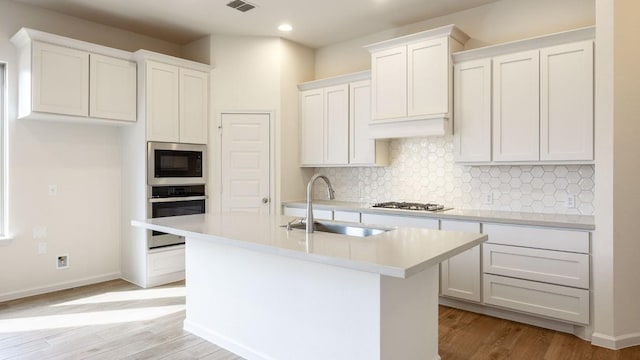 kitchen with sink, appliances with stainless steel finishes, white cabinets, a kitchen island with sink, and backsplash