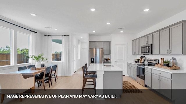 kitchen with stainless steel appliances, gray cabinets, an island with sink, and light wood-type flooring