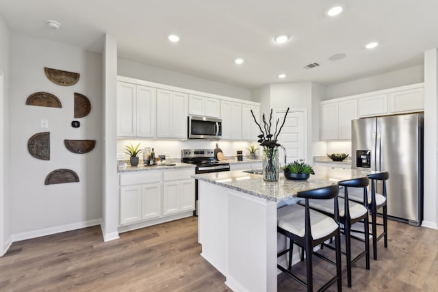 kitchen with sink, a kitchen island with sink, white cabinetry, stainless steel appliances, and light stone counters