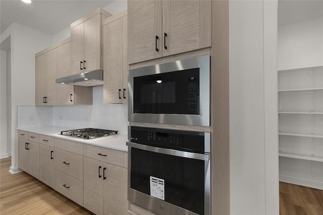 kitchen with under cabinet range hood, light countertops, light wood-type flooring, decorative backsplash, and appliances with stainless steel finishes