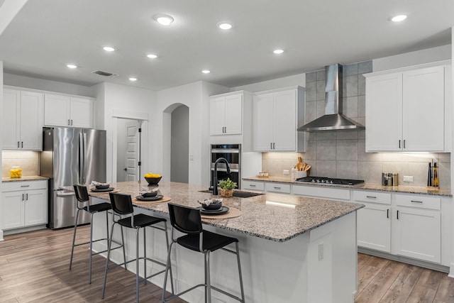 kitchen with a center island with sink, white cabinetry, appliances with stainless steel finishes, and wall chimney range hood