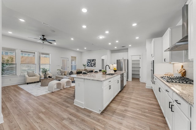 kitchen featuring appliances with stainless steel finishes, an island with sink, sink, white cabinets, and wall chimney range hood