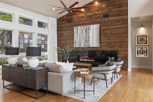 living room with ceiling fan, high vaulted ceiling, light hardwood / wood-style floors, and wood walls