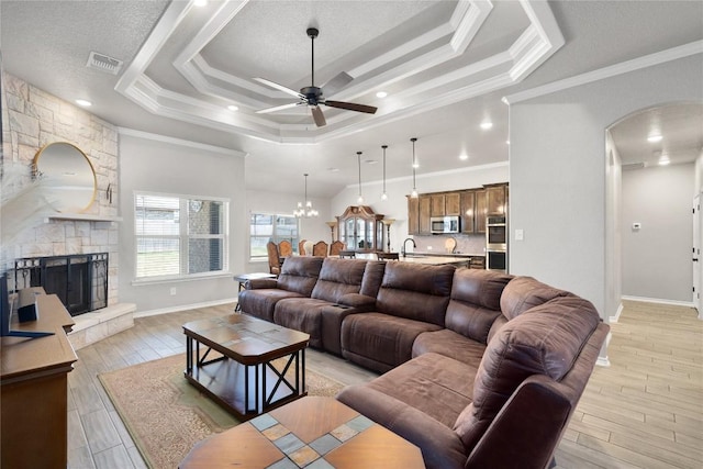 living room with ornamental molding, a stone fireplace, a raised ceiling, and ceiling fan with notable chandelier