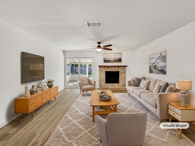 living area with light wood-type flooring, a ceiling fan, a stone fireplace, and baseboards