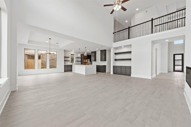 unfurnished living room with a tray ceiling, ceiling fan with notable chandelier, light hardwood / wood-style flooring, and a high ceiling