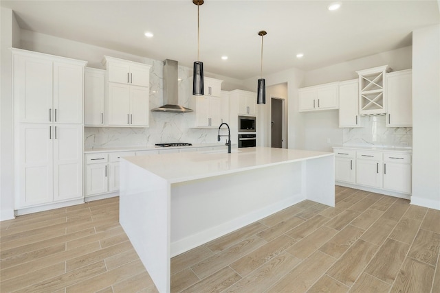 kitchen with wood finish floors, a sink, appliances with stainless steel finishes, wall chimney exhaust hood, and a center island with sink