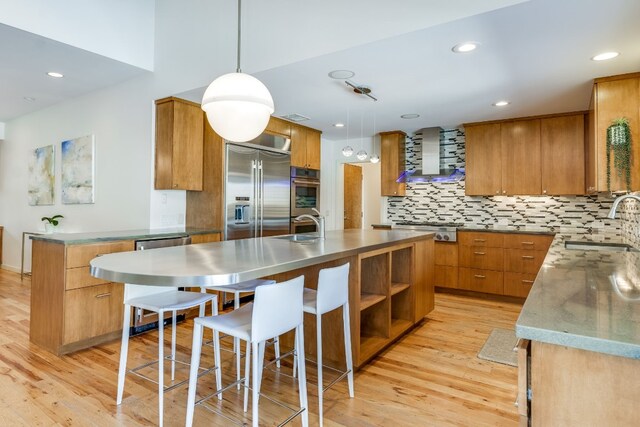 kitchen featuring sink, wall chimney range hood, appliances with stainless steel finishes, hanging light fixtures, and an island with sink