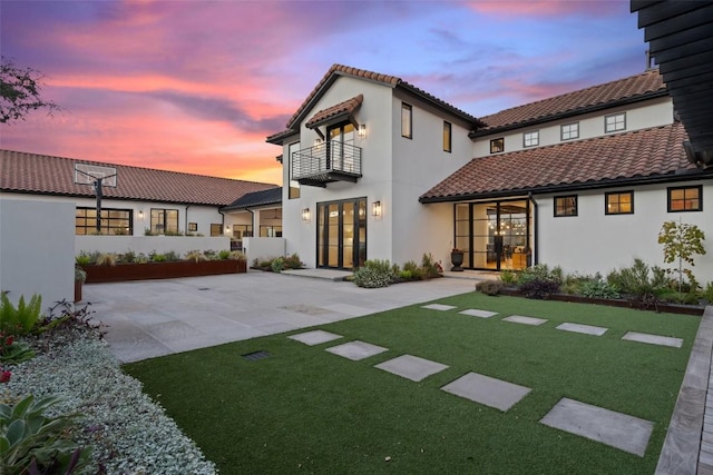 back house at dusk featuring a balcony, a patio, and a lawn