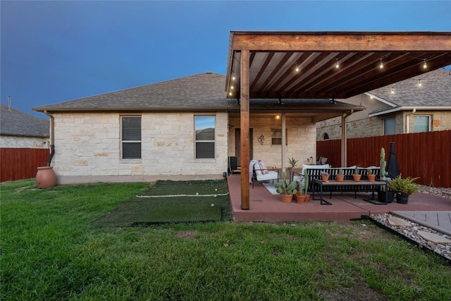 rear view of property with a lawn, stone siding, fence, a shingled roof, and a patio area