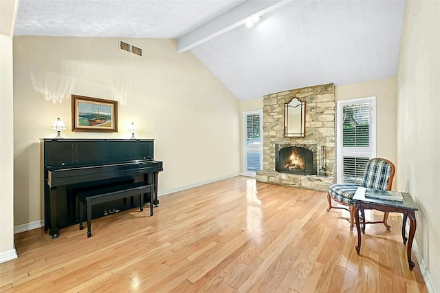 living area with beam ceiling, high vaulted ceiling, light hardwood / wood-style floors, a textured ceiling, and a stone fireplace