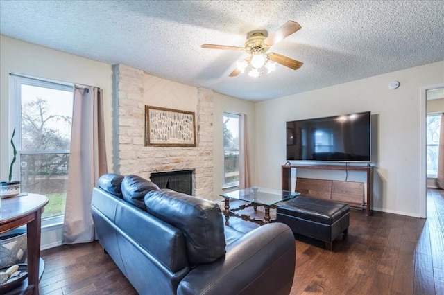 living room featuring baseboards, dark wood finished floors, a ceiling fan, a textured ceiling, and a stone fireplace