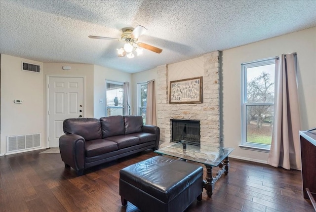 living room featuring ceiling fan, a stone fireplace, dark wood-type flooring, and a textured ceiling