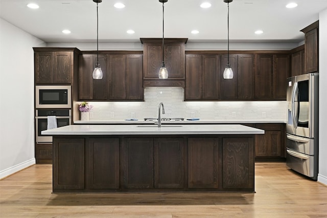 kitchen with decorative light fixtures, dark brown cabinets, light wood-type flooring, stainless steel appliances, and a kitchen island with sink