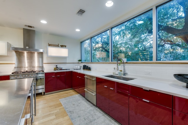 kitchen with sink, backsplash, stainless steel gas range oven, light wood-type flooring, and wall chimney exhaust hood